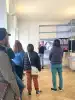 Foto: In Rückenansicht schauen Besucher*innen  auf einen männlichen Ausstellungs-Guide. Er gebärdet in Deutscher Gebärden Sprache und informiert zur Ausstellung. Im Hintergrund ist ein Metallregal mit Monitoren, das Teil einer Kunstinstallation ist.