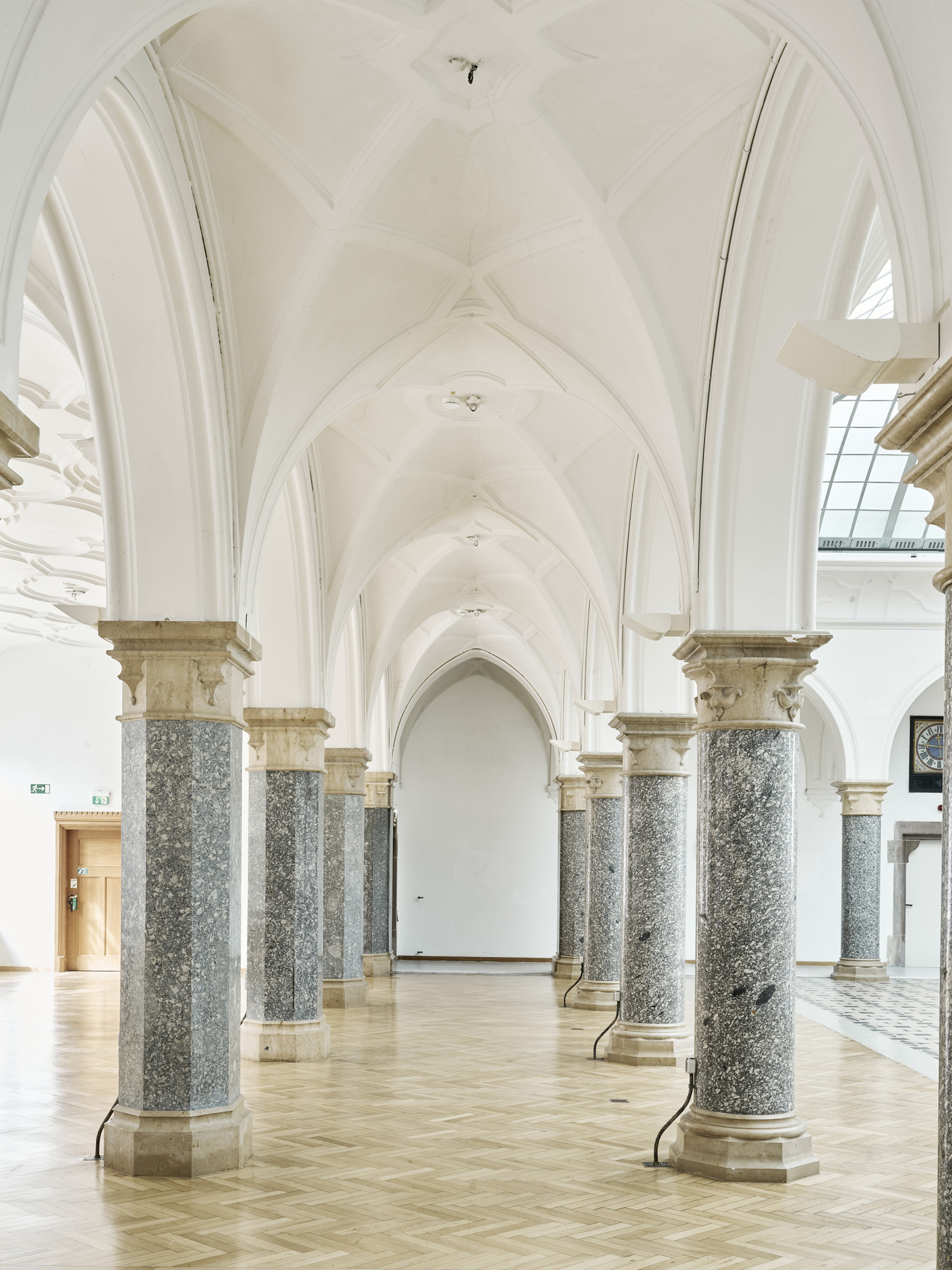 Foto: Ein Gang mit weißem Deckengewölbe führt zu einer weißen Wand am Ende des Gangs. Auf beiden Seiten wird das Gewölbe von jeweils 4 grauen Granitpfeilern getragen.