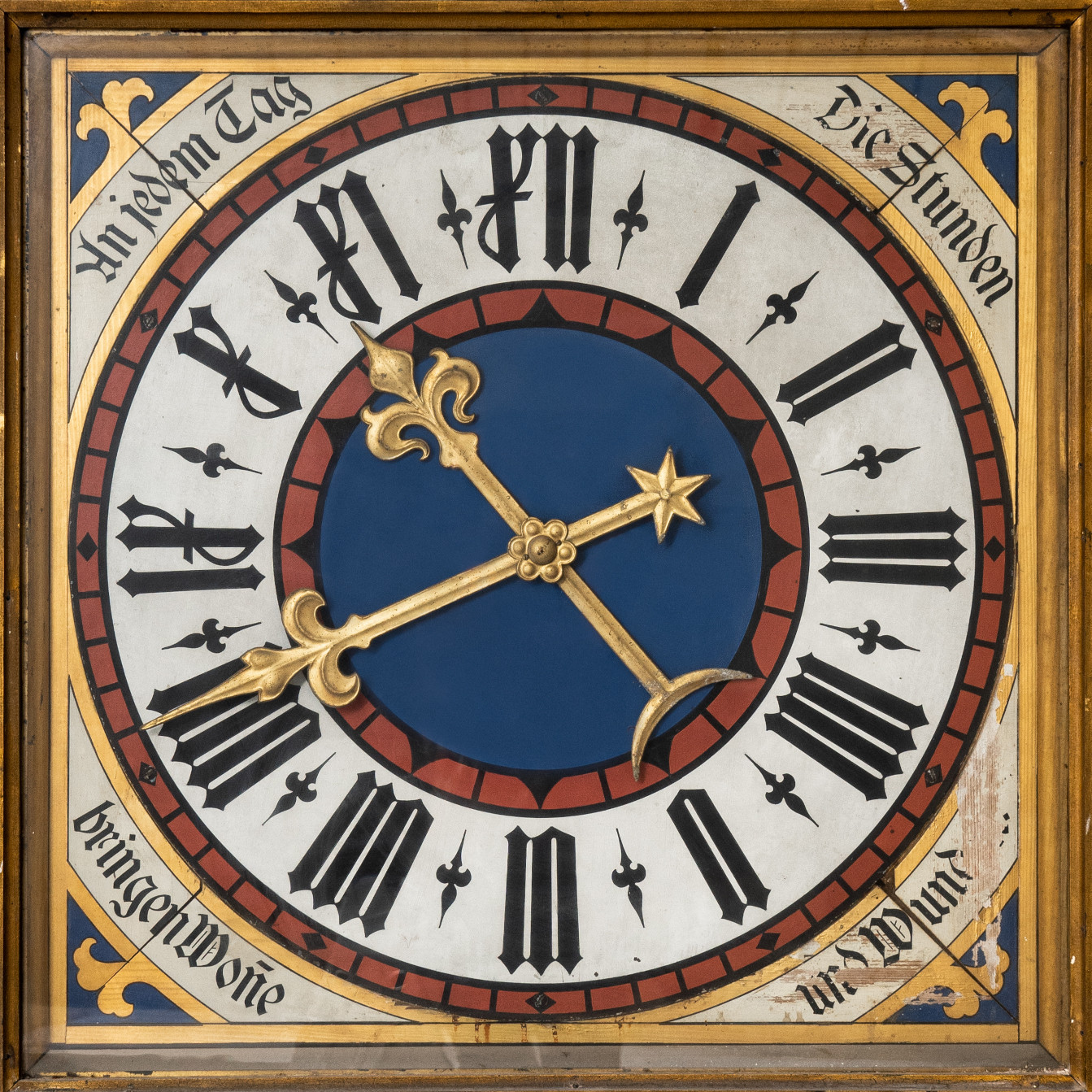 Historische Uhr mit goldenen verzierten Zeigern. Das Ziffernblatt ist in den Farben rot, weiß, schwarz und blau gehalten. Ein Schriftzug in historischer Schrift umrandet das Ziffernblatt mit den Worten: An jedem Tag  die Stunden bringen Wonne und Wunden