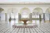 Foto: Der rötliche Marmorbrunnen steht im Zentrum der Halle auf einem schwarz-weißen Fliesenfußboden. Dahinter öffnen fünf Torbögen mit grauen Granitpfeilern den Durchgang zu einem weiteren Raum. Transparente hellgelbe und rosafarbene Vorhänge hängen zwis